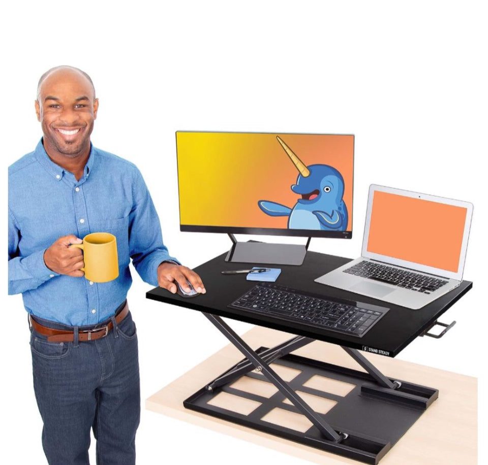 Standing desk setup with Stand Steady X-Elite Pro converter; spacious surface holds monitor, laptop, and keyboard; user standing with coffee mug.