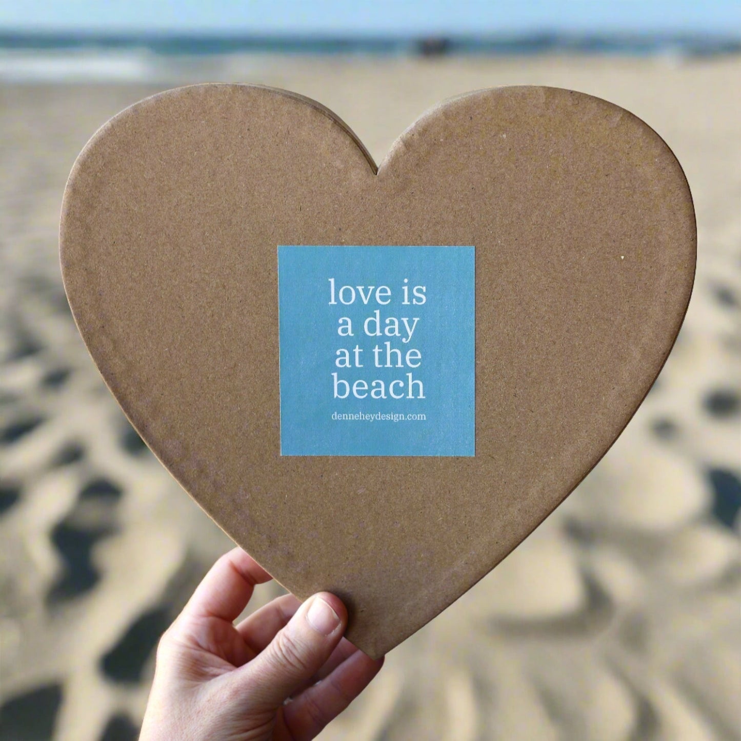 Heart-shaped beach mystery box with "love is a day at the beach" label held on sandy beach background