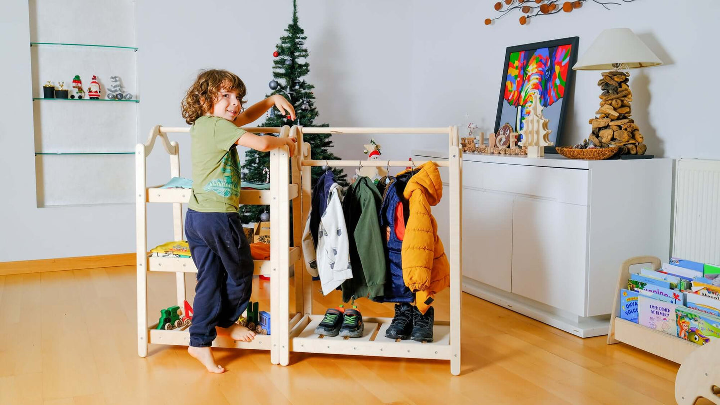 Child interacting with Montessori wooden wardrobe and clothes rack featuring hanging jackets and shelves for organized toddler clothing in a bright room.