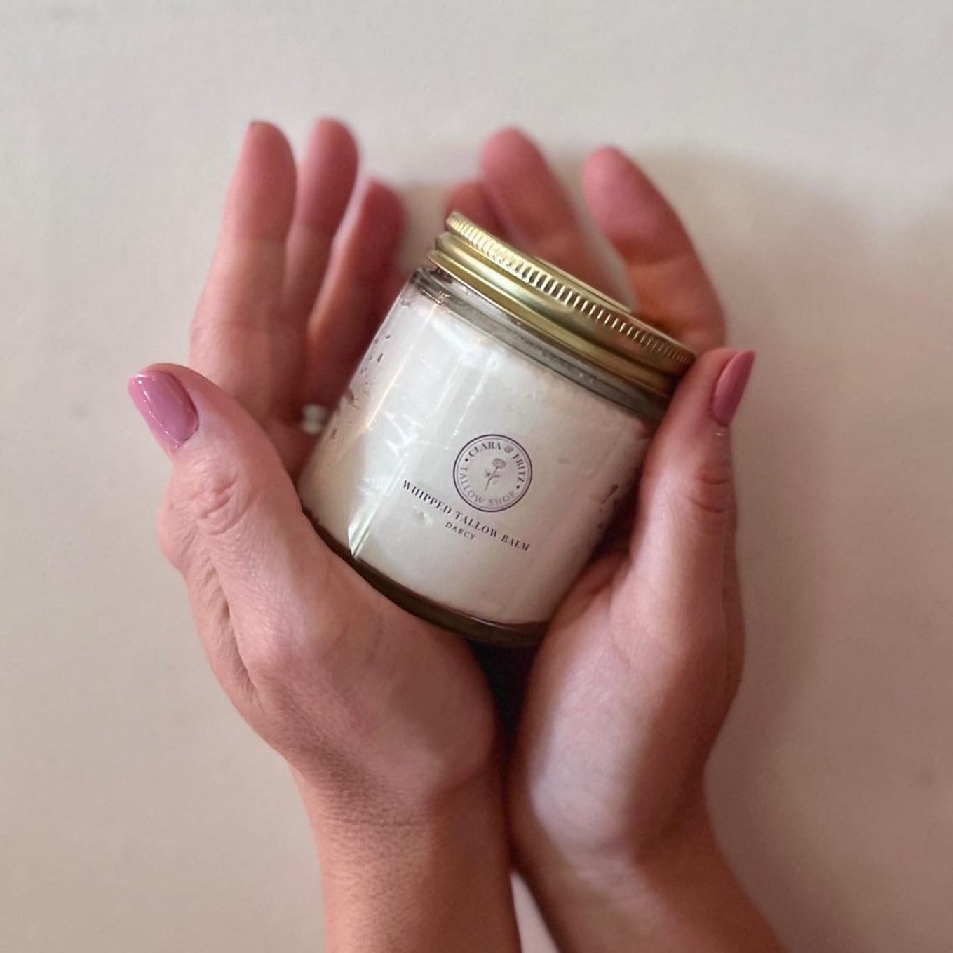 Jar of Darcy Frankincense & Cedarwood scented whipped tallow balm held between hands with pink nail polish
