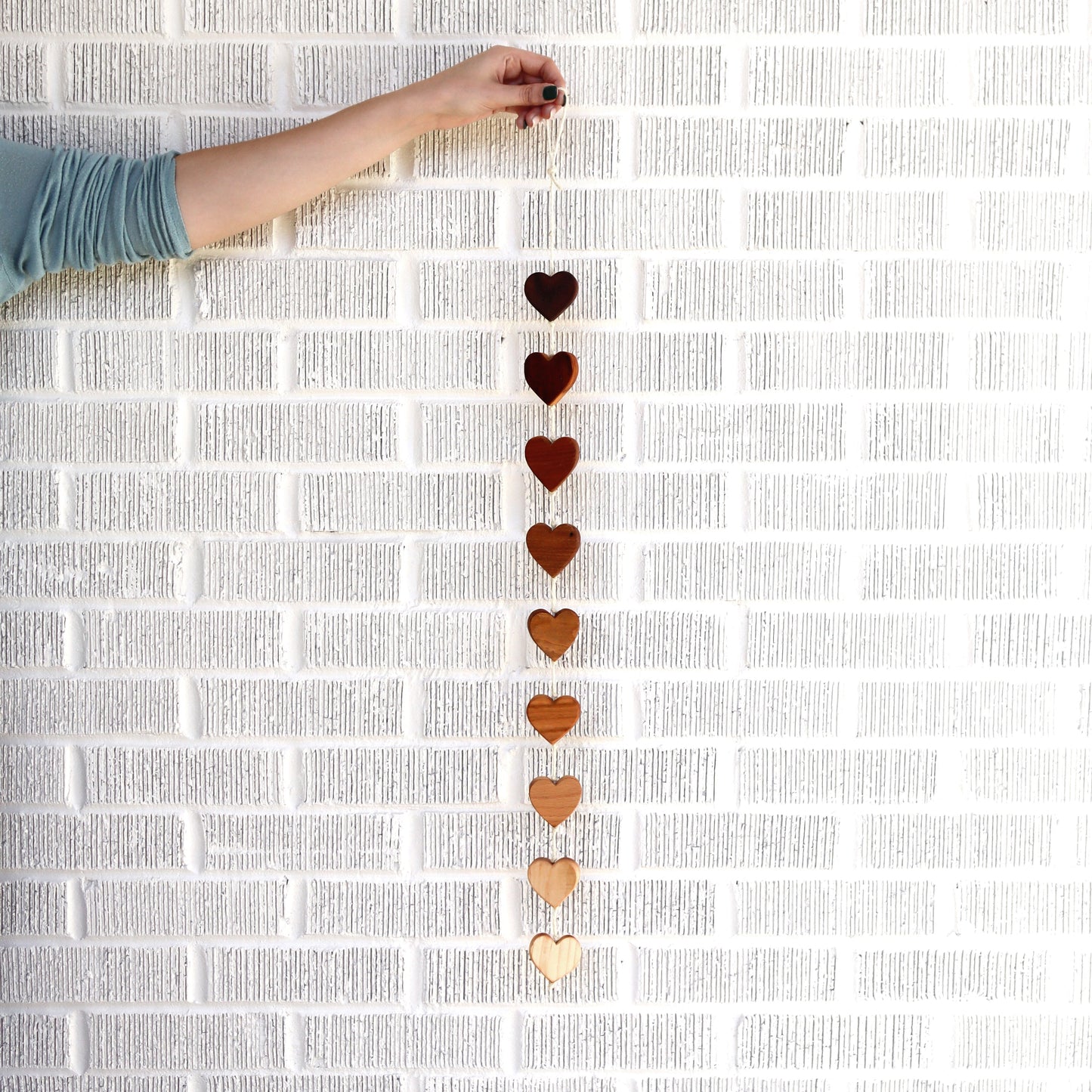Handcrafted Ombré Heartstrings wall decor with nine connected wooden hearts in varying natural hardwood shades hanging against a white brick wall.