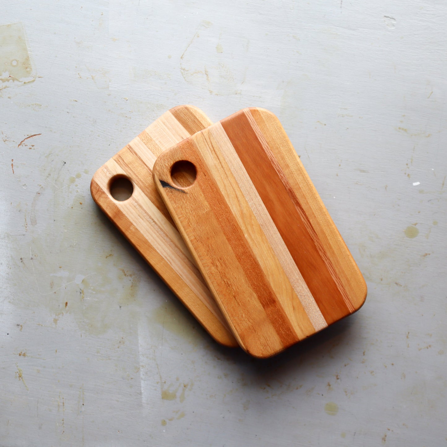 Two handmade laminated hardwood cheese boards with contrasting light and dark stripes and rounded corners on a light surface