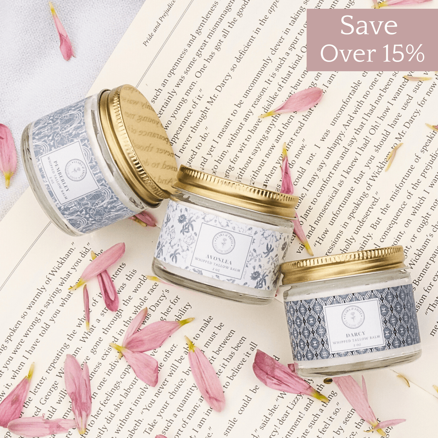 Signature Scent Trio of whipped tallow balms with lavender, chamomile, frankincense, cedarwood, and orange scents in glass jars with gold lids and floral labels on an open book with pink flower petals and a save over 15% tag.