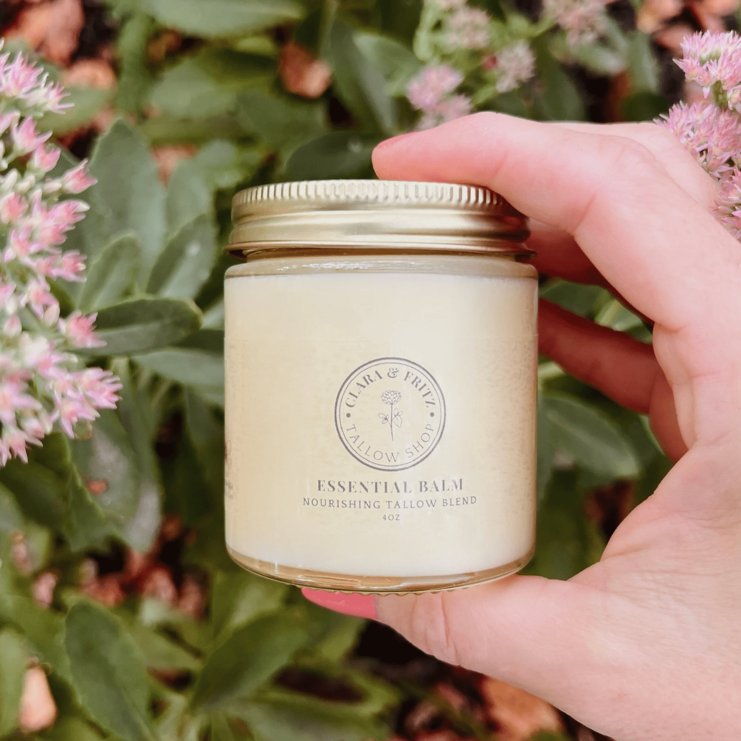 Jar of Clara & Fritz Essential Balm nourishing tallow blend held outdoors near green plants and pink flowers.