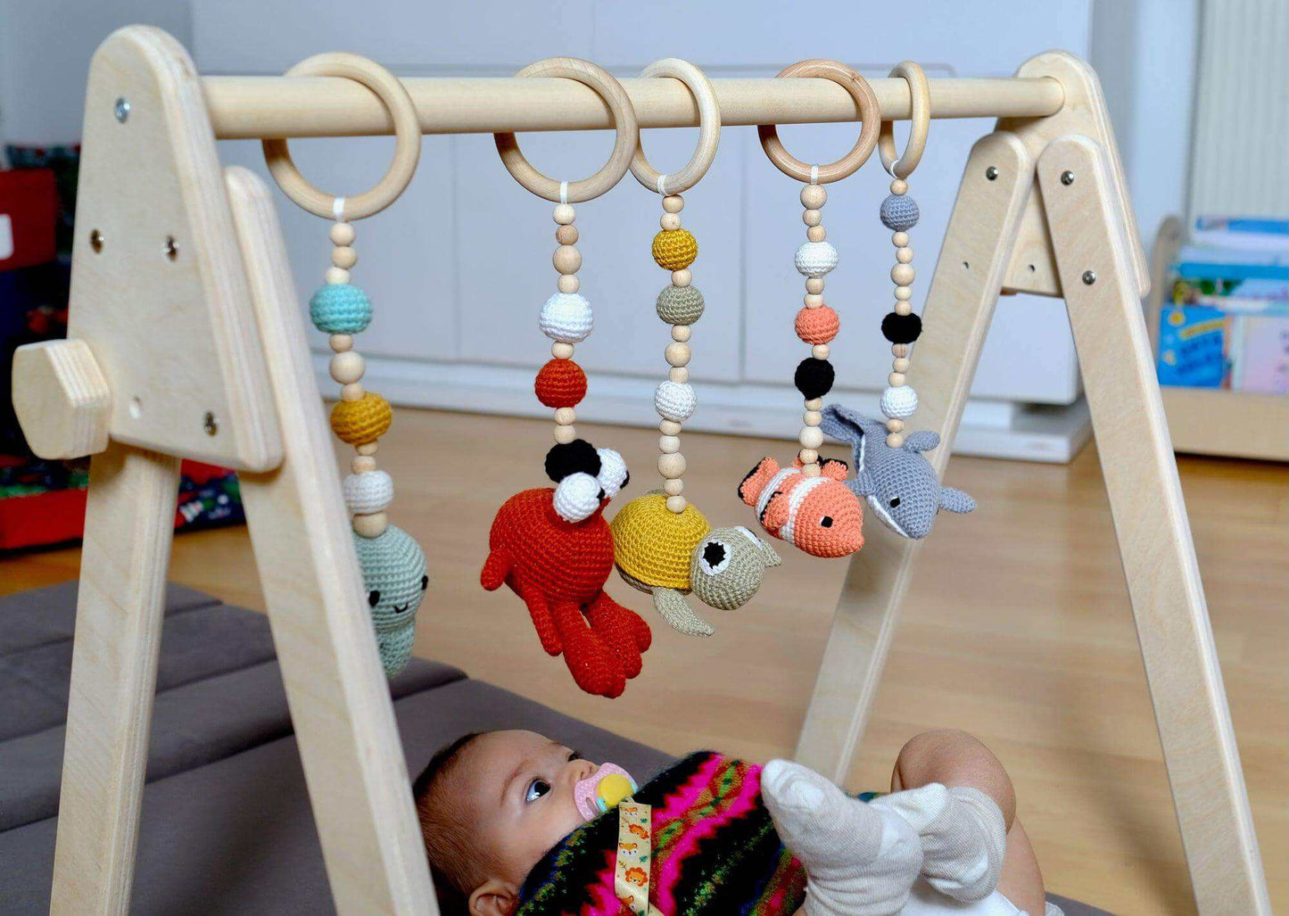 Montessori baby gym with wooden frame and five colorful crochet hanging toys over a baby lying underneath.