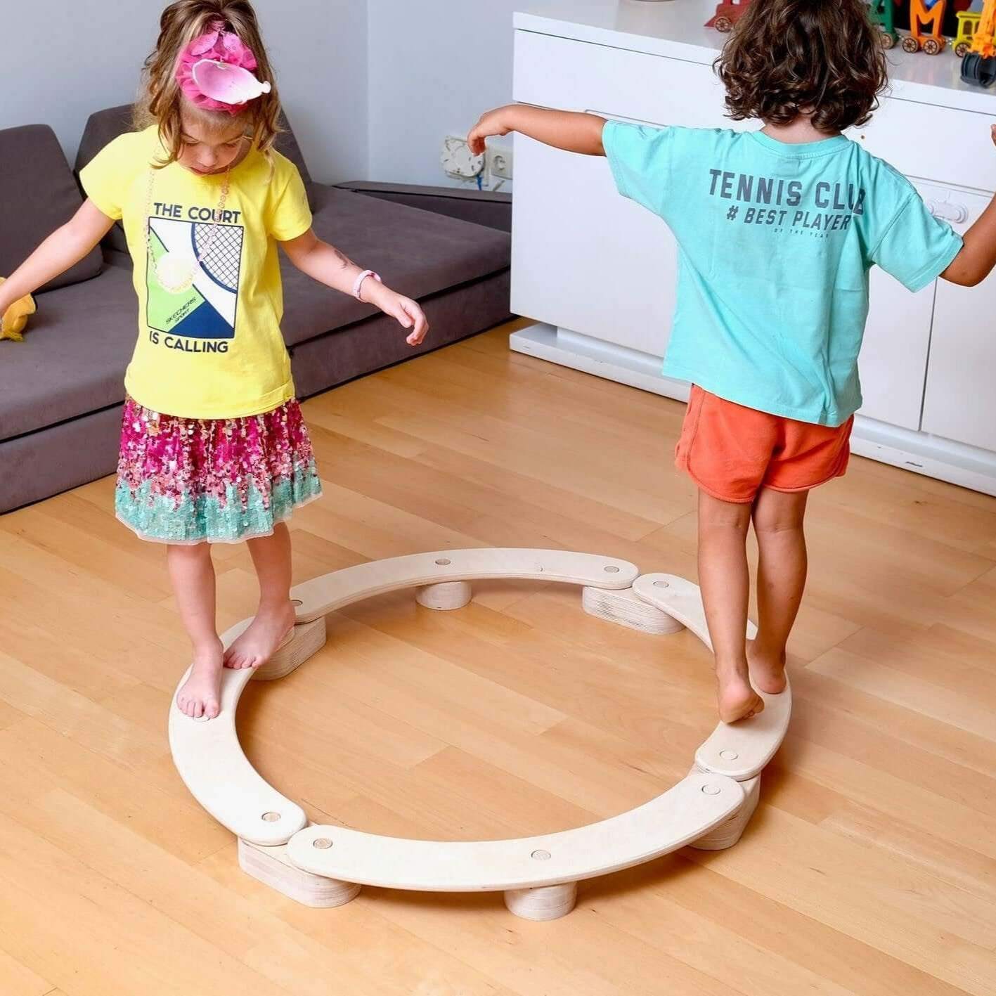 Children balancing on a circular wooden balance beam set on a wooden floor in a playroom
