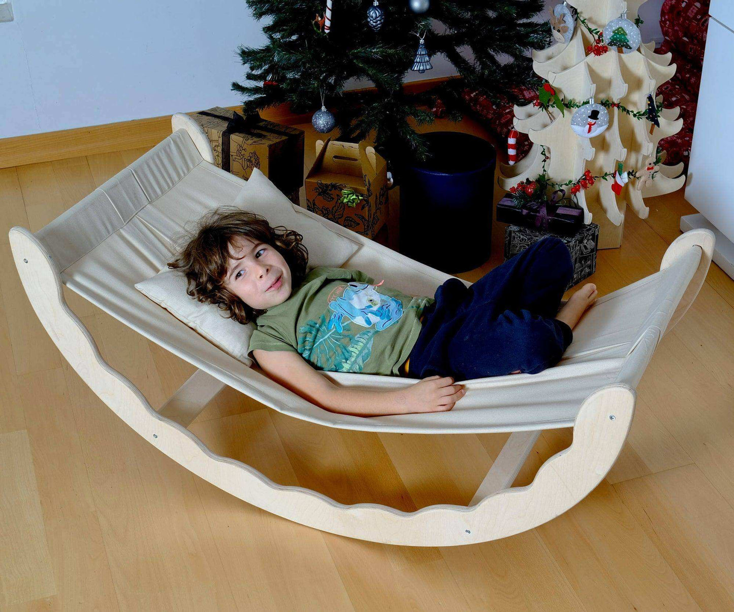 Toddler relaxing on a wooden hammock with fabric sling indoors near a decorated Christmas tree