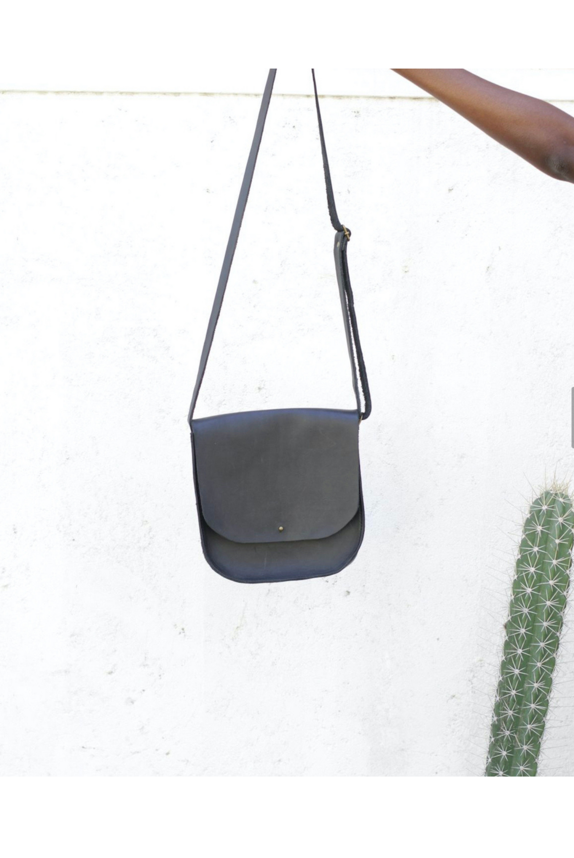 Black full-grain leather saddlebag with adjustable crossbody strap and flap closure hanging against white wall.