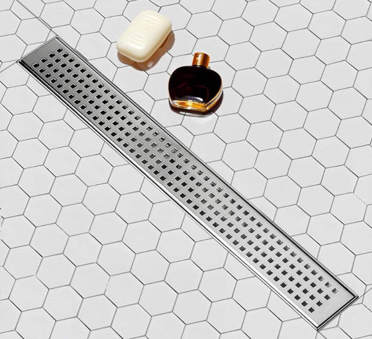 SaniteModar Linear Shower Drain 60 inch with removable square hole panel in brushed 304 stainless steel installed on hexagonal white tile shower floor with soap and perfume bottle nearby