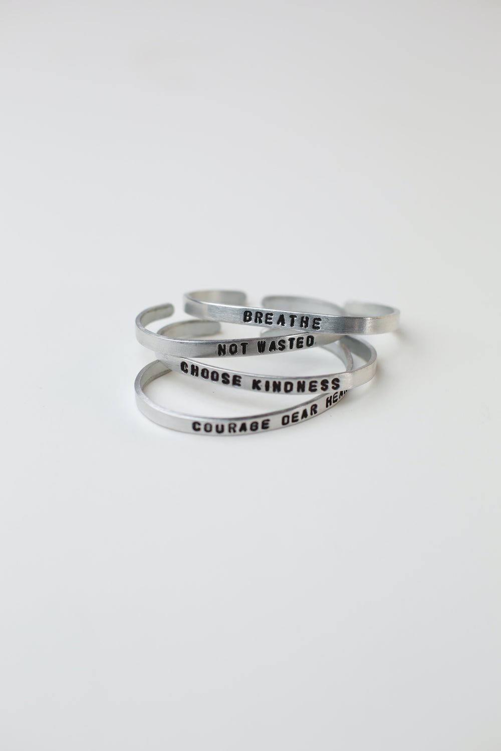Set of upcycled aluminum story cuffs engraved with inspirational phrases like "Breathe," "Not Wasted," "Choose Kindness," and "Courage Dear Heart"