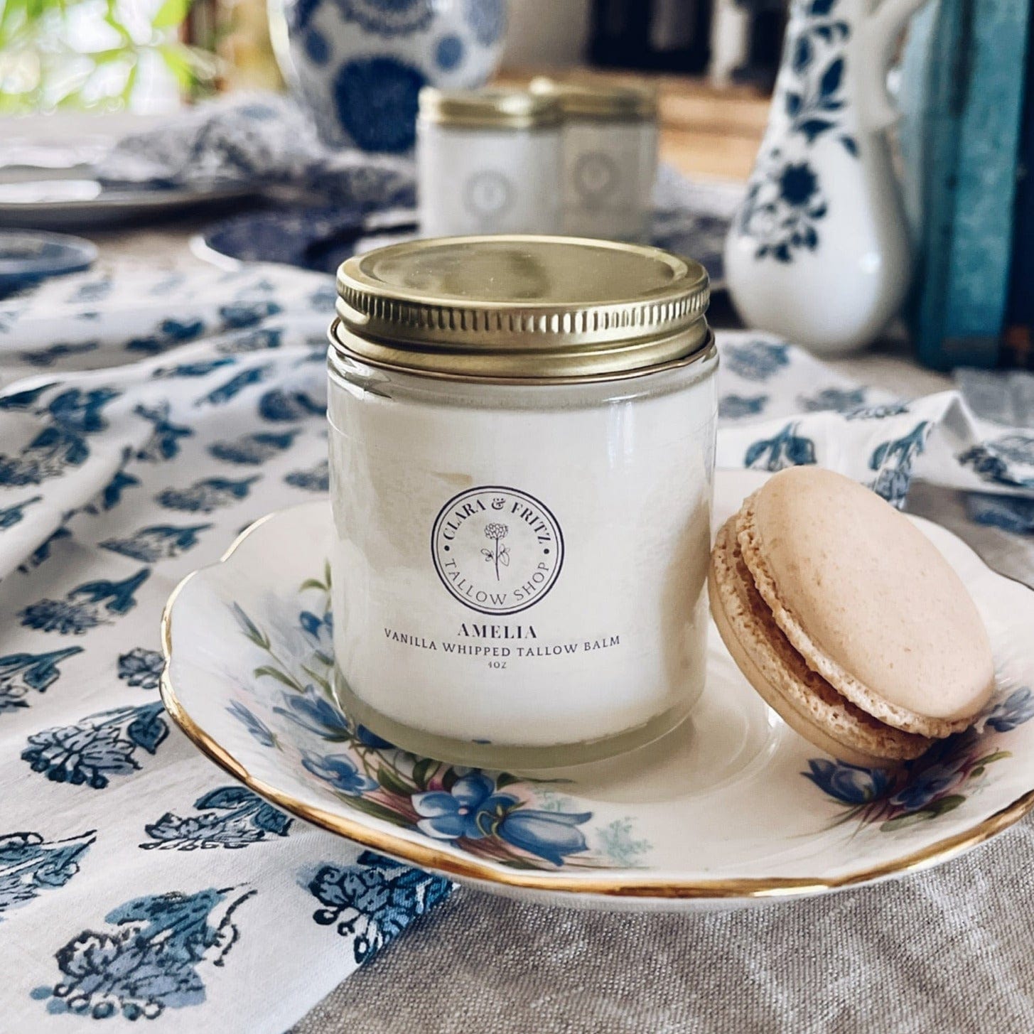 Amelia Vanilla Whipped Tallow Balm in glass jar with gold lid on floral plate next to vanilla macaron