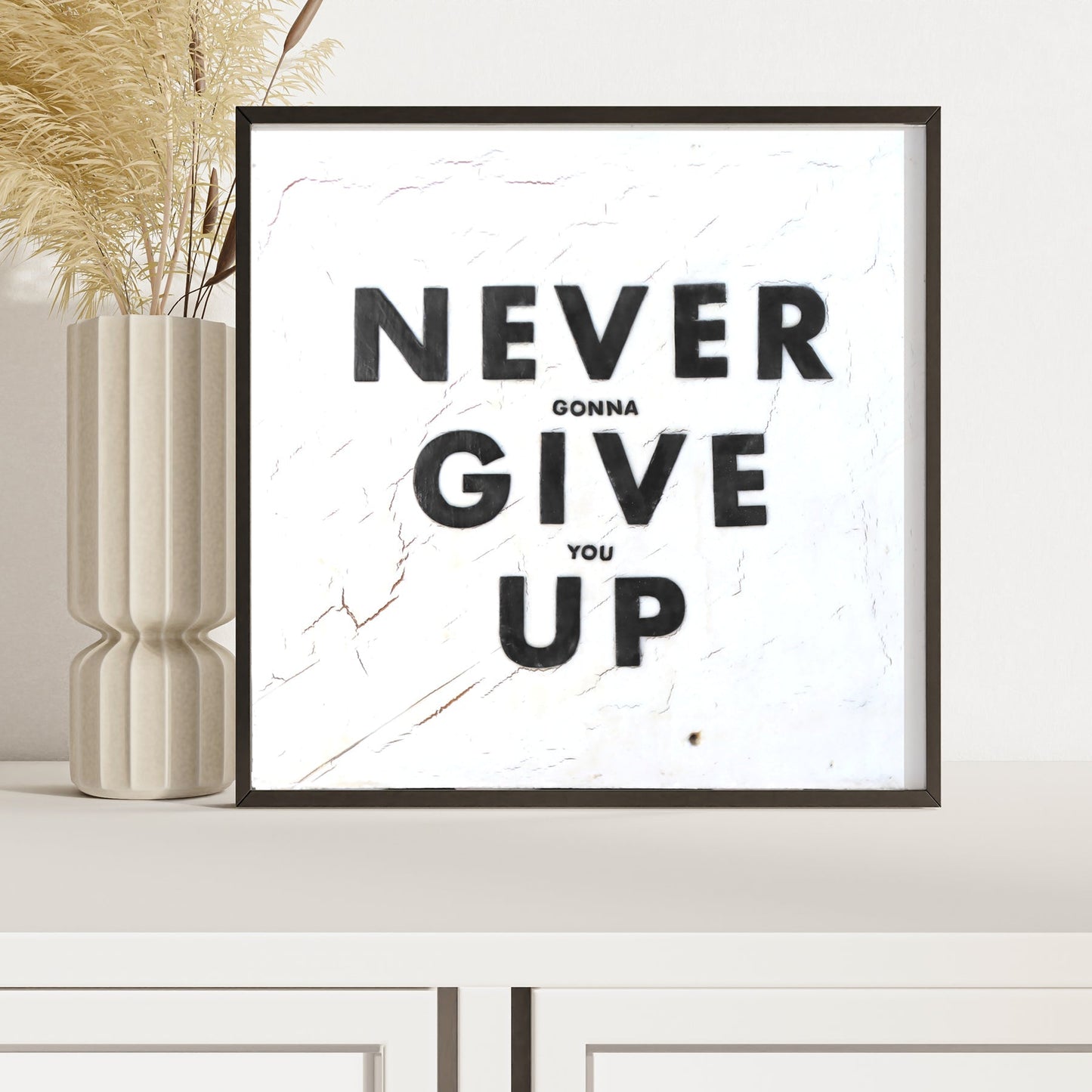 Framed Never Give Up art print with bold black text on white background displayed on white shelf next to beige ceramic vase with pampas grass