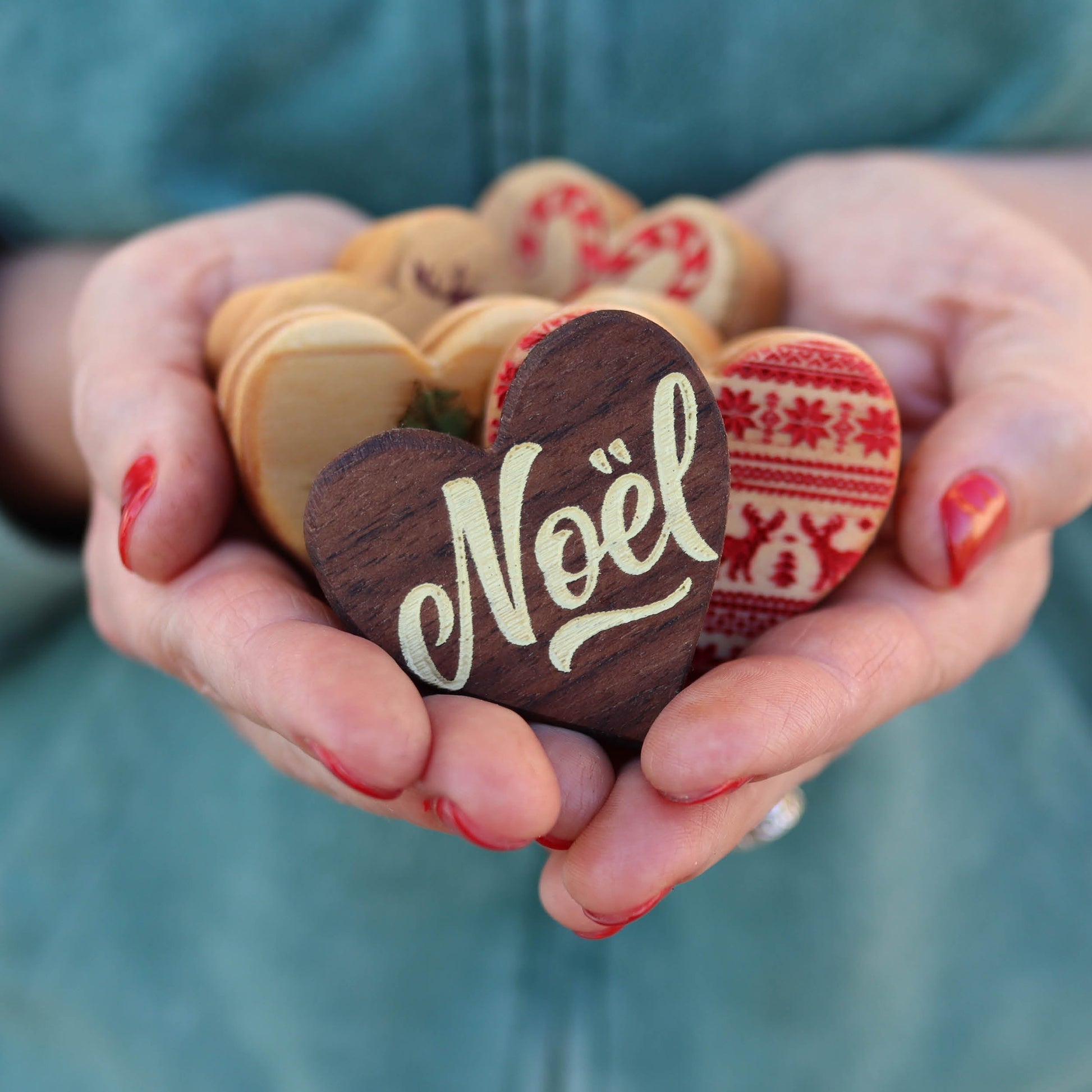 Person holding a variety of handmade wooden heart ornaments including one with "Noël" written on it in festive Christmas designs