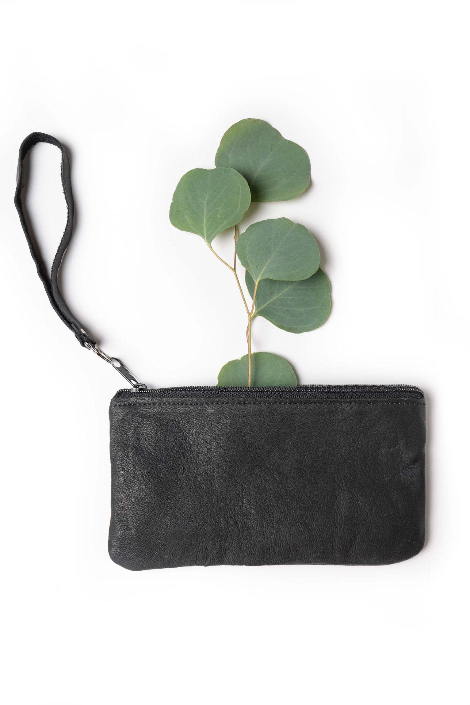 Black handmade leather pouch with wristlet strap and zipper closure on white background with green leaves