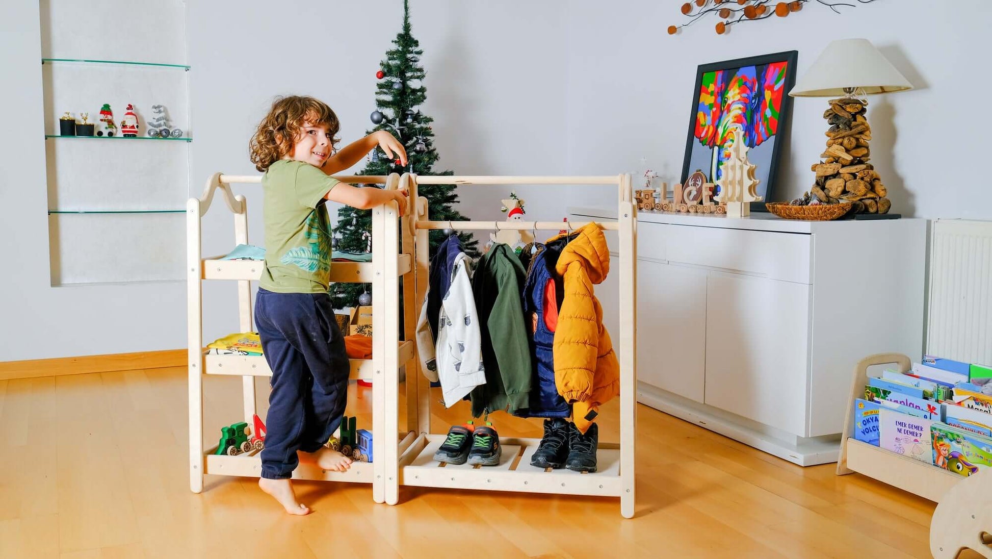 Child interacting with Montessori wooden wardrobe and clothes rack featuring hanging jackets and shelves for organized toddler clothing in a bright room.