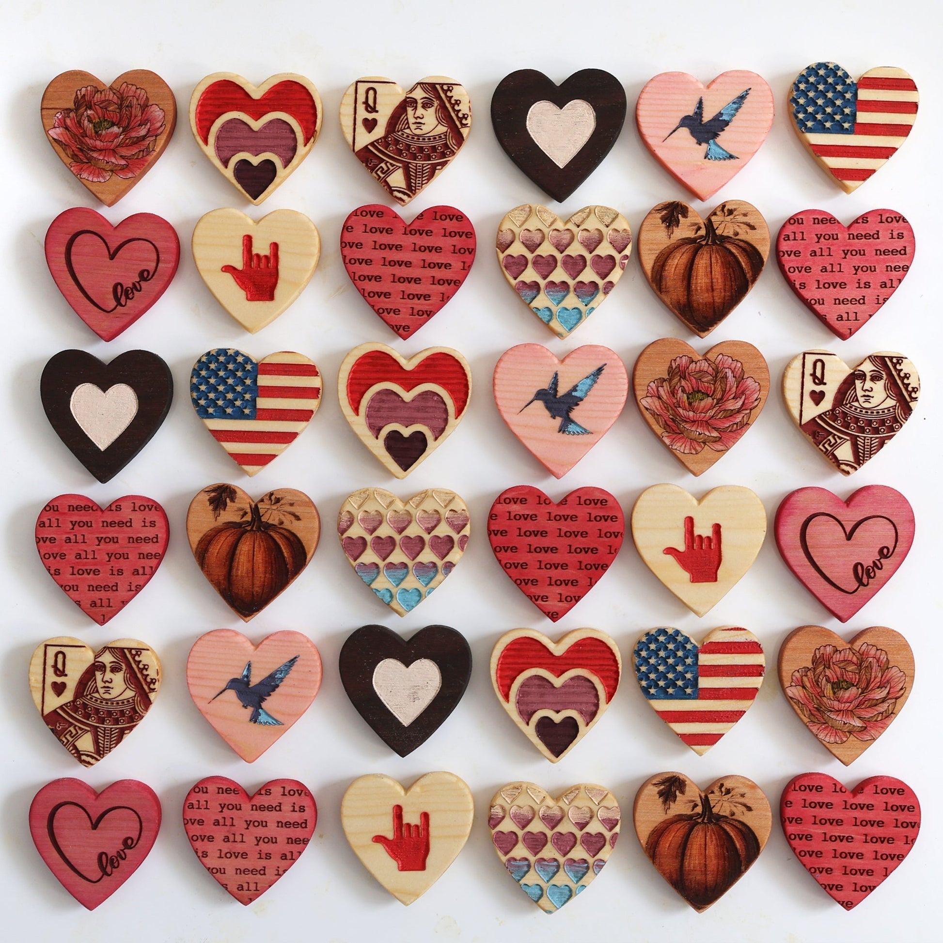 Assorted handmade engraved heart magnets made from reclaimed wood featuring various designs including love text, hearts, pumpkins, birds, and flag patterns.