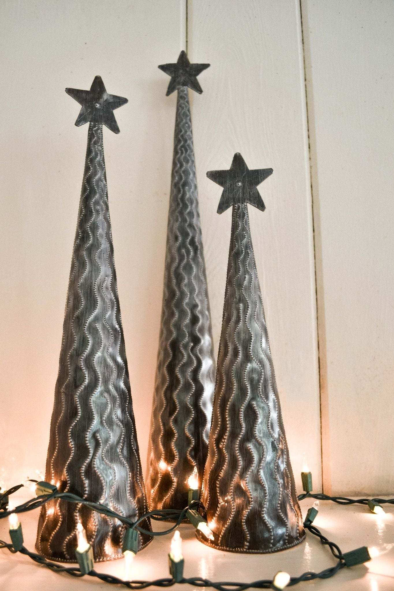 Set of three standing metal trees made from recycled steel with star toppers and textured patterns, illuminated by string lights.