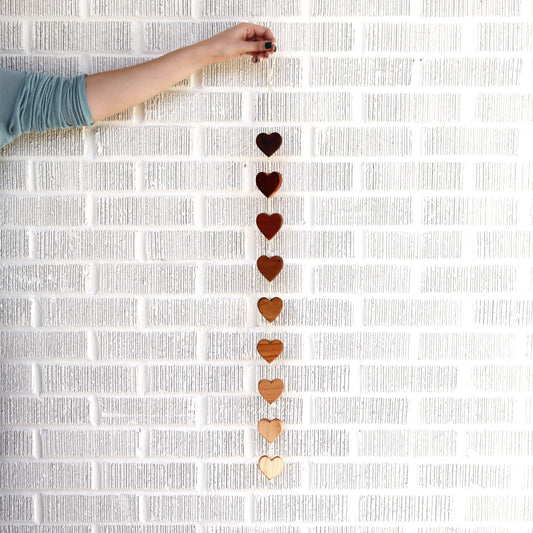 Handcrafted Ombré Heartstrings wall decor with nine connected wooden hearts in varying natural hardwood shades hanging against a white brick wall.