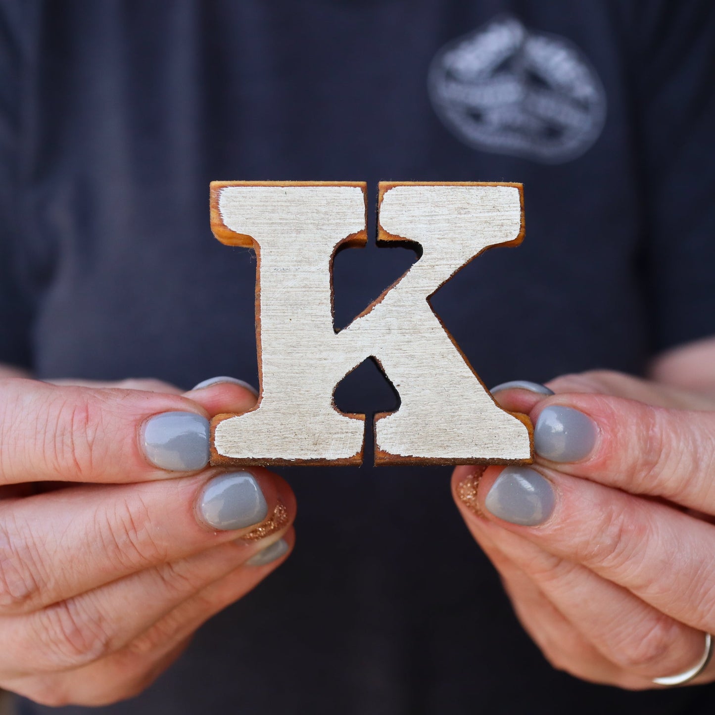 Handcrafted little wooden letter K made from reclaimed wood held between fingers with gray nail polish
