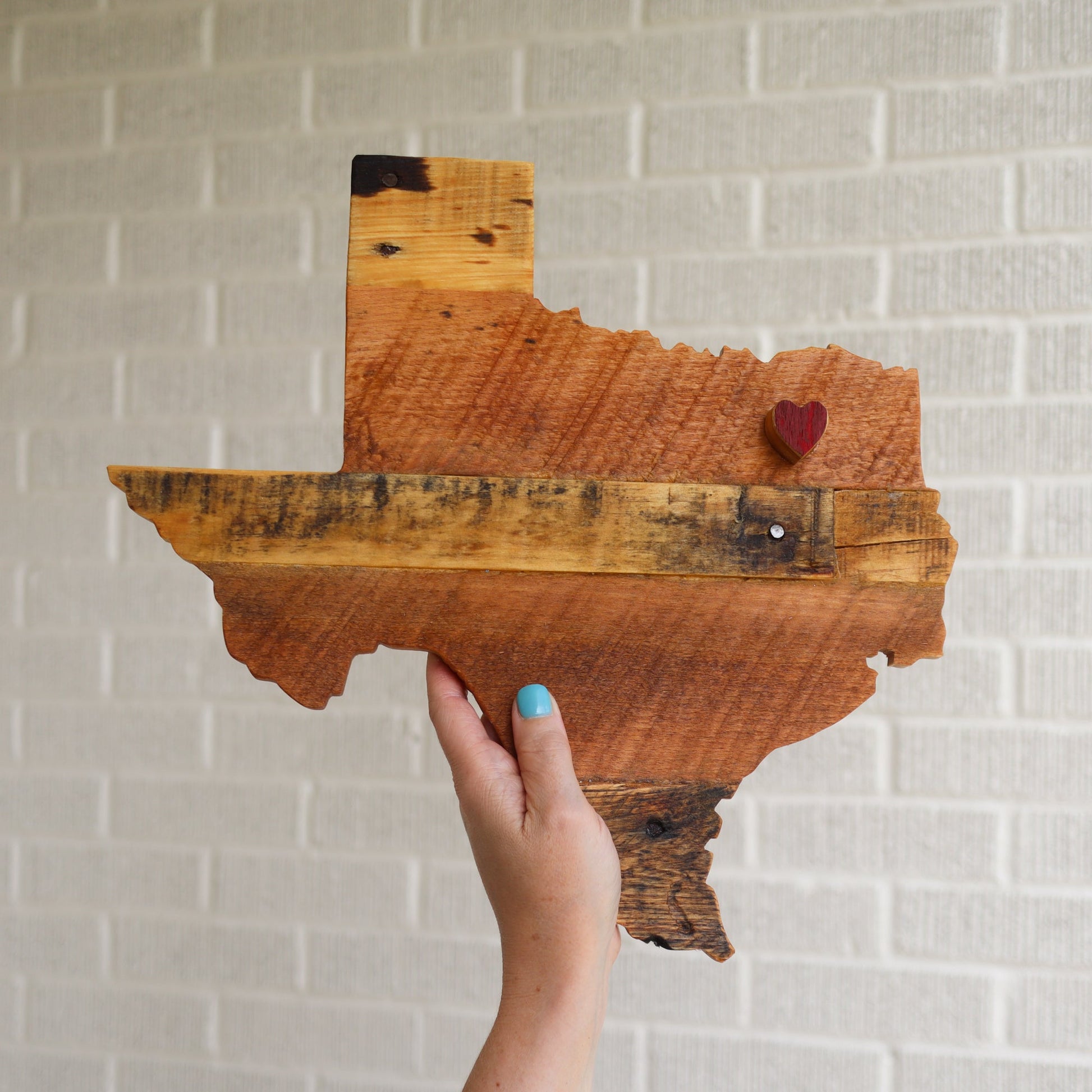 Handcrafted pallet wood state art shaped like Texas with a small red heart marker, held against a white brick wall background