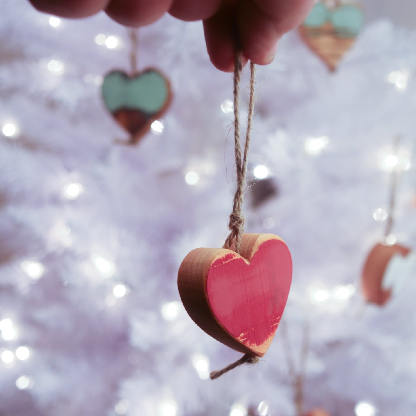 Handmade reclaimed wooden heart ornament with jute twine hanger against a blurred white Christmas tree background with lights and similar ornaments