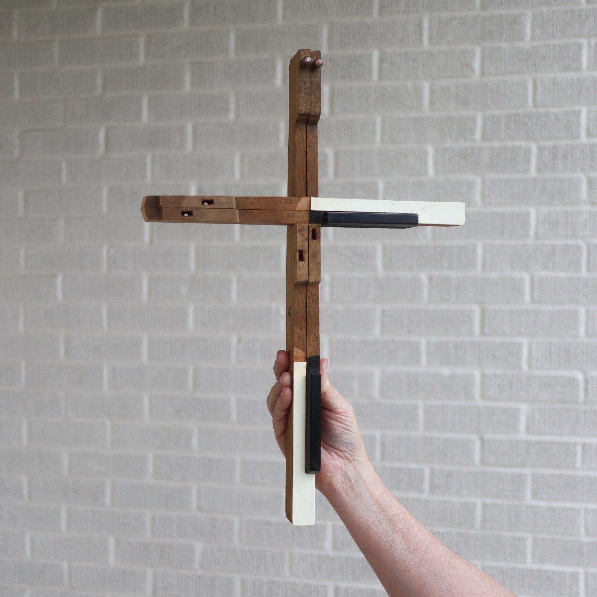 Handmade piano key cross crafted from reclaimed antique piano keys with natural wood aging, held against a gray brick wall background