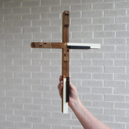 Handmade piano key cross crafted from reclaimed antique piano keys with natural wood aging, held against a gray brick wall background