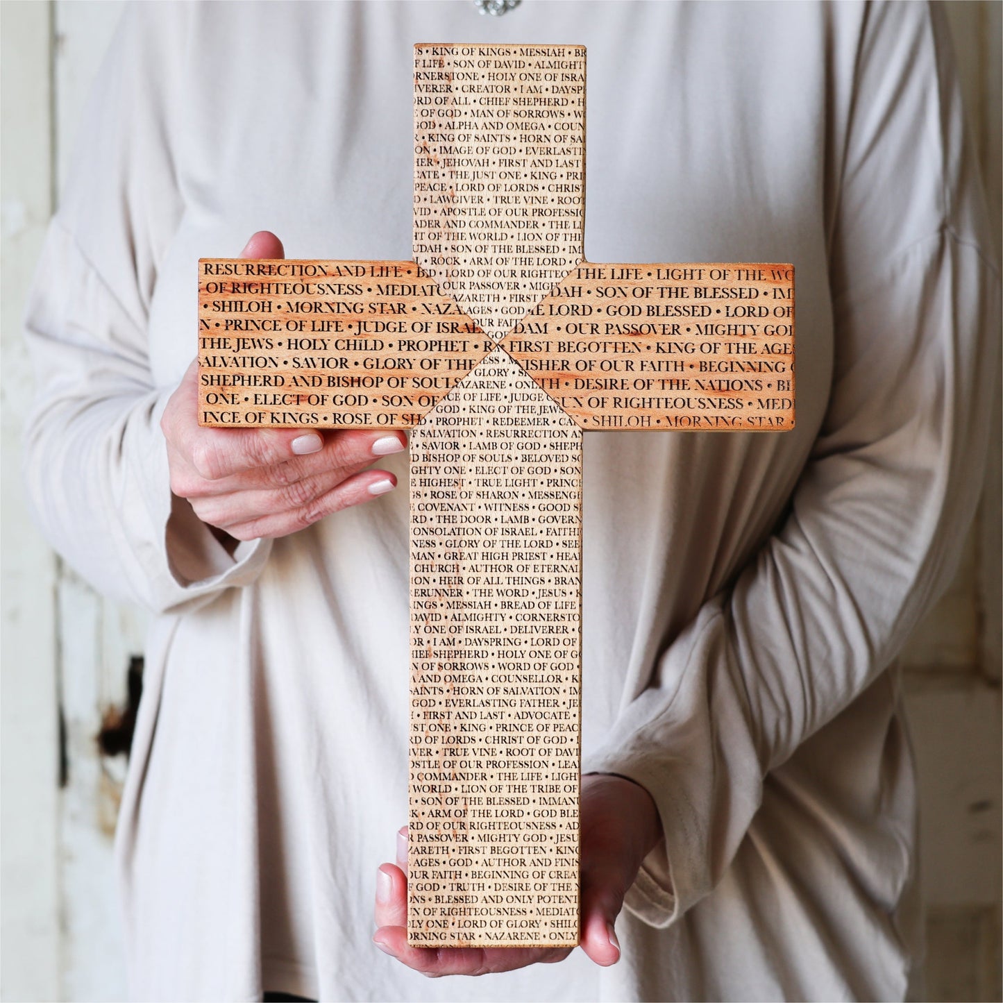 Handcrafted Names of the Savior Cross made from reclaimed piano wood with engraved Jesus names, held by person in beige robe