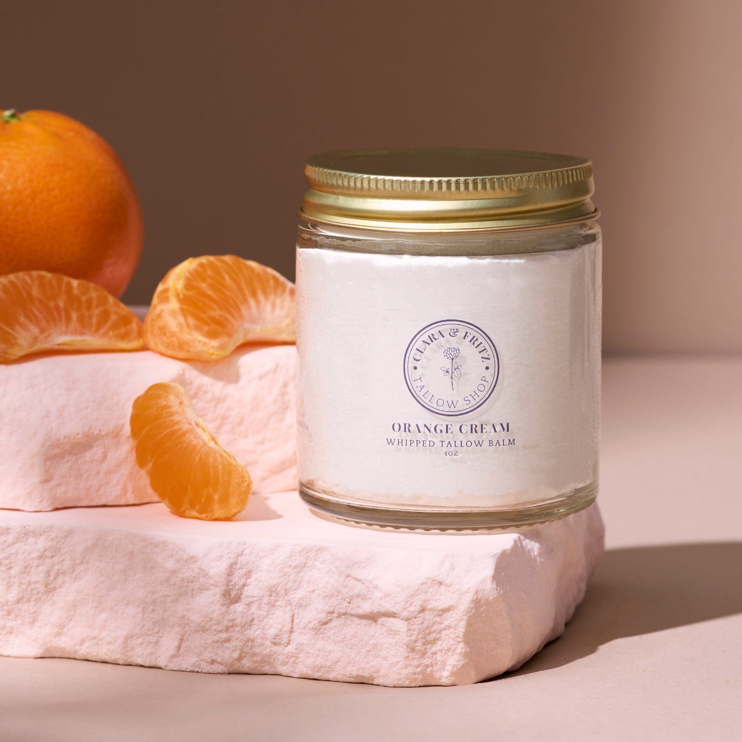 Jar of Orange Cream Whipped Tallow Balm beside fresh orange slices on a pale pink stone pedestal