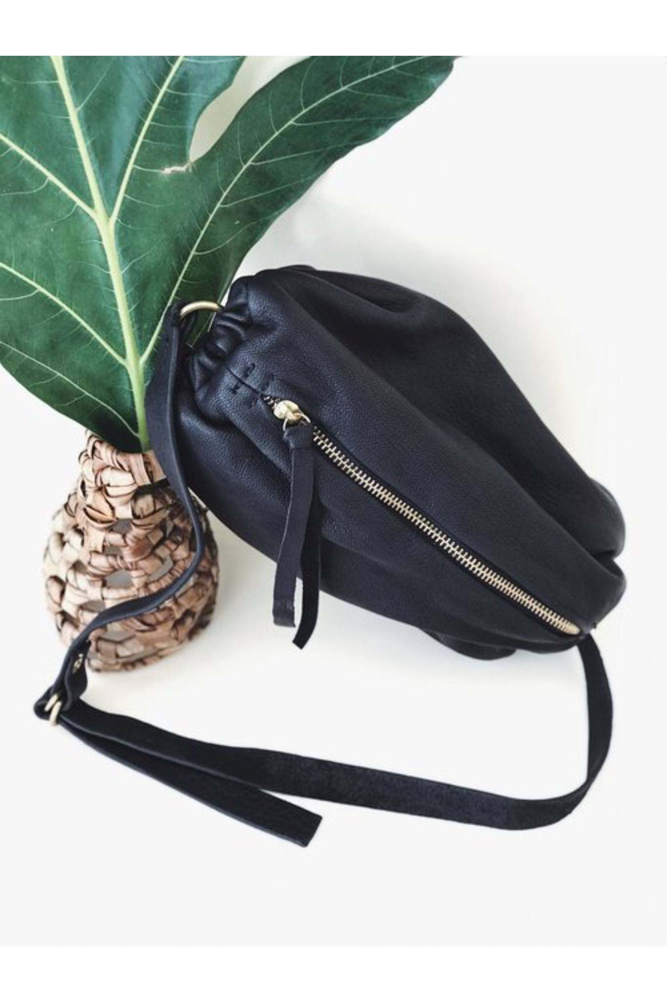 Black leather scrunchie bag with zipper and adjustable strap placed beside a woven vase with green leaf.