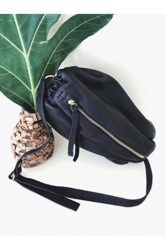 Black leather scrunchie bag with zipper and adjustable strap placed beside a woven vase with green leaf.