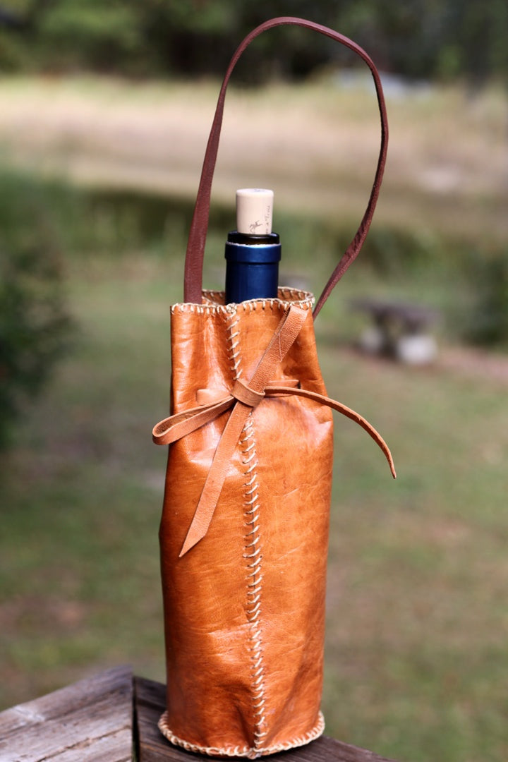 Handmade leather bottle holder with stitched detailing and shoulder strap carrying a wine bottle outdoors