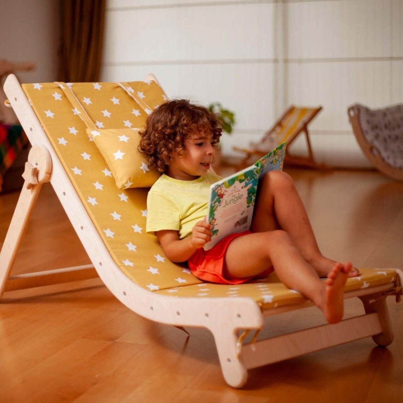 Child sitting on adjustable lounge chair with yellow star-patterned comfy pad reading a book indoors