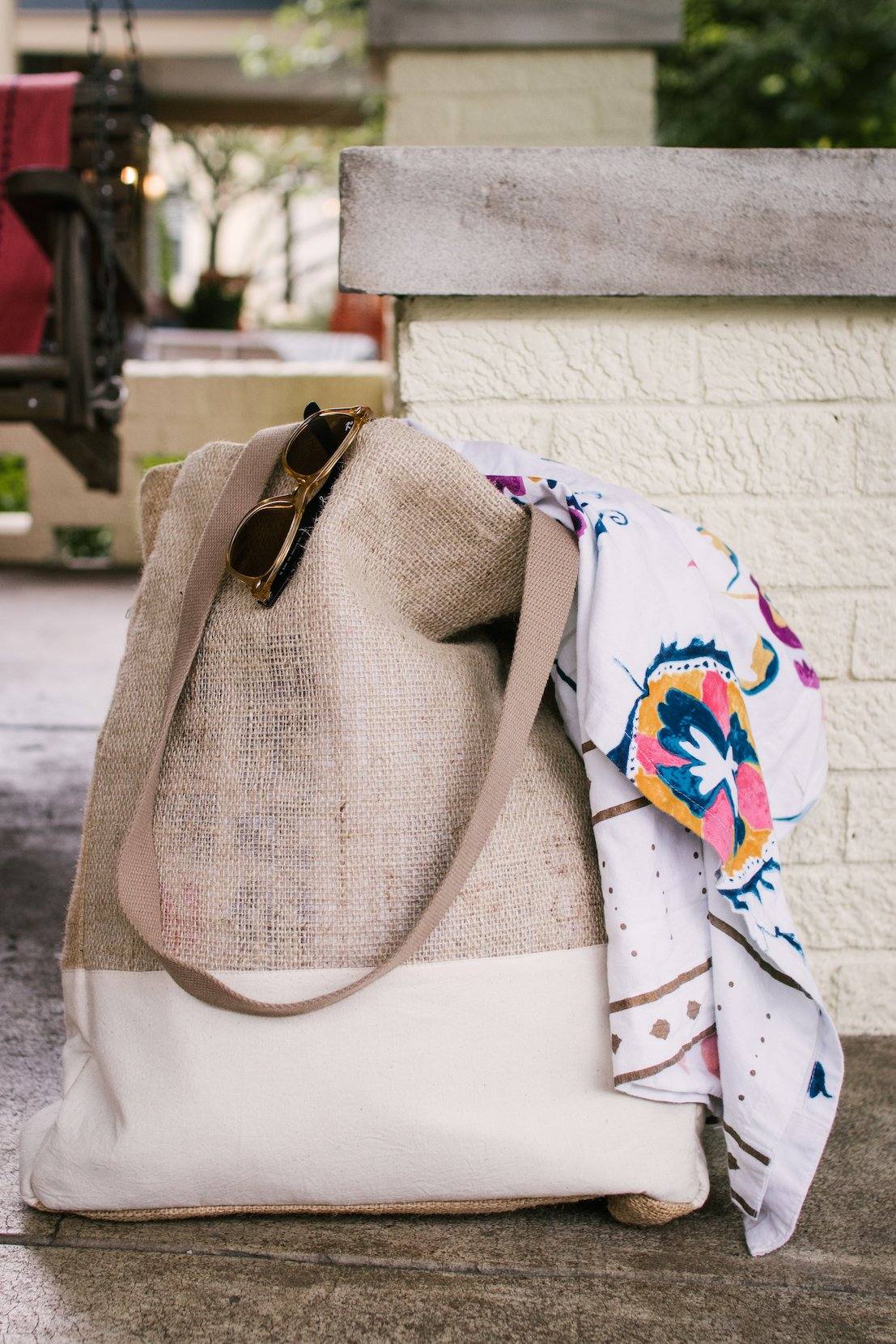 Burlap Market Tote bag with tan straps, made from recycled burlap and local materials, featuring a white bottom and carrying sunglasses and a colorful cloth.