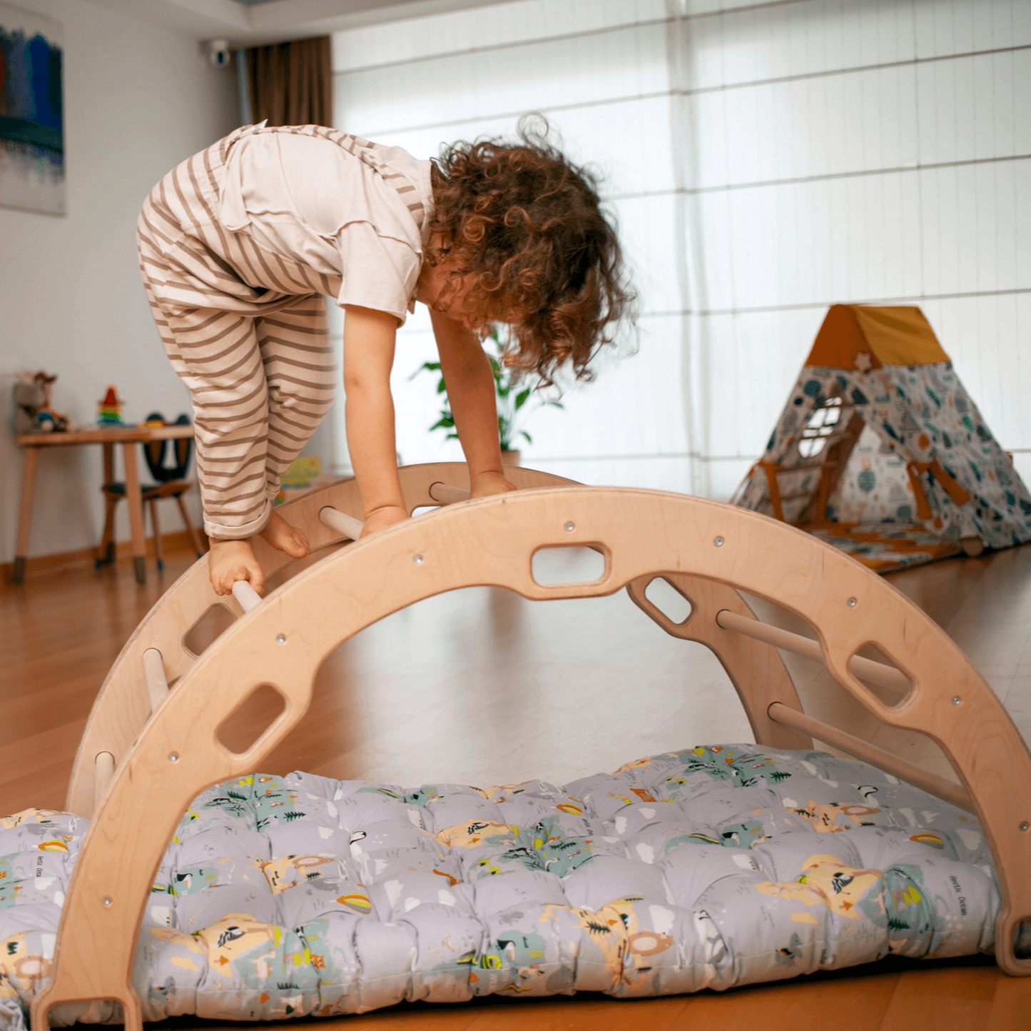 Child climbing on large Montessori Climbing Arch made of natural wood with patterned pillow set underneath in a bright playroom.