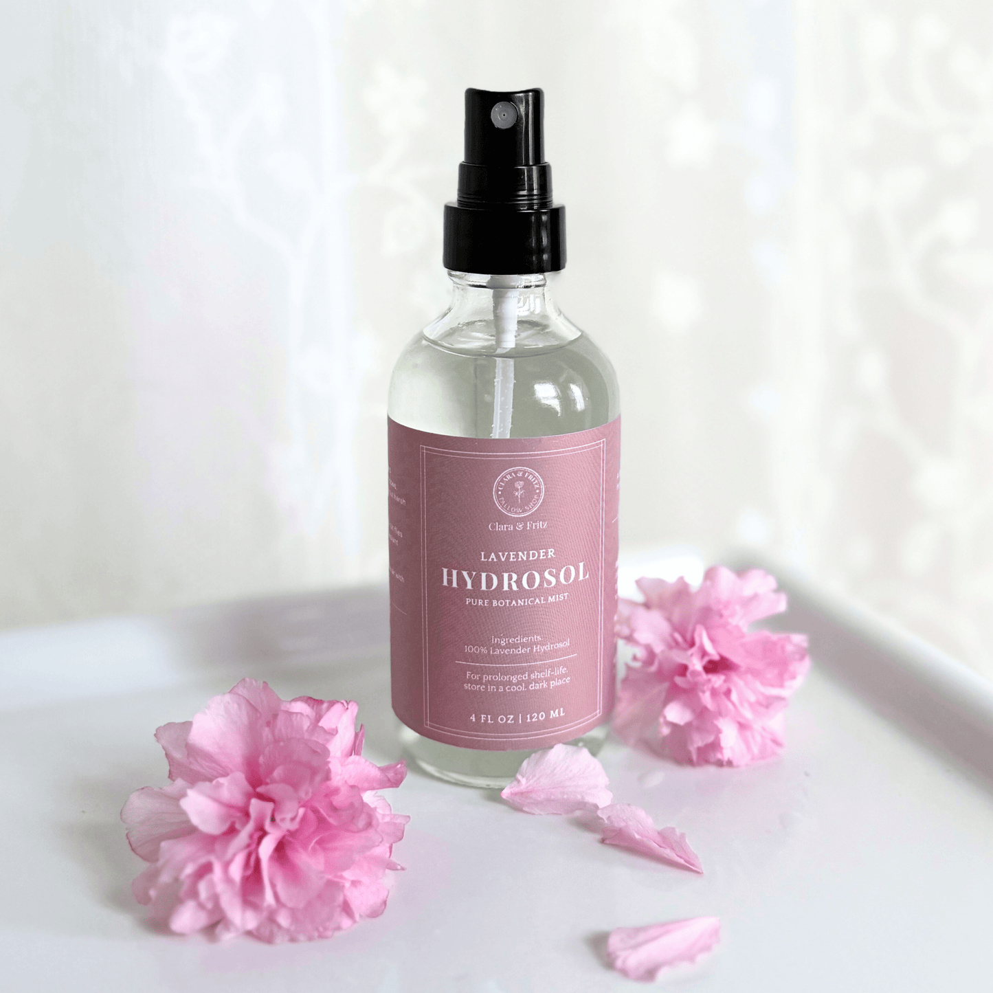 Clear spray bottle of lavender hydrosol with pink label on white surface surrounded by pink flower petals