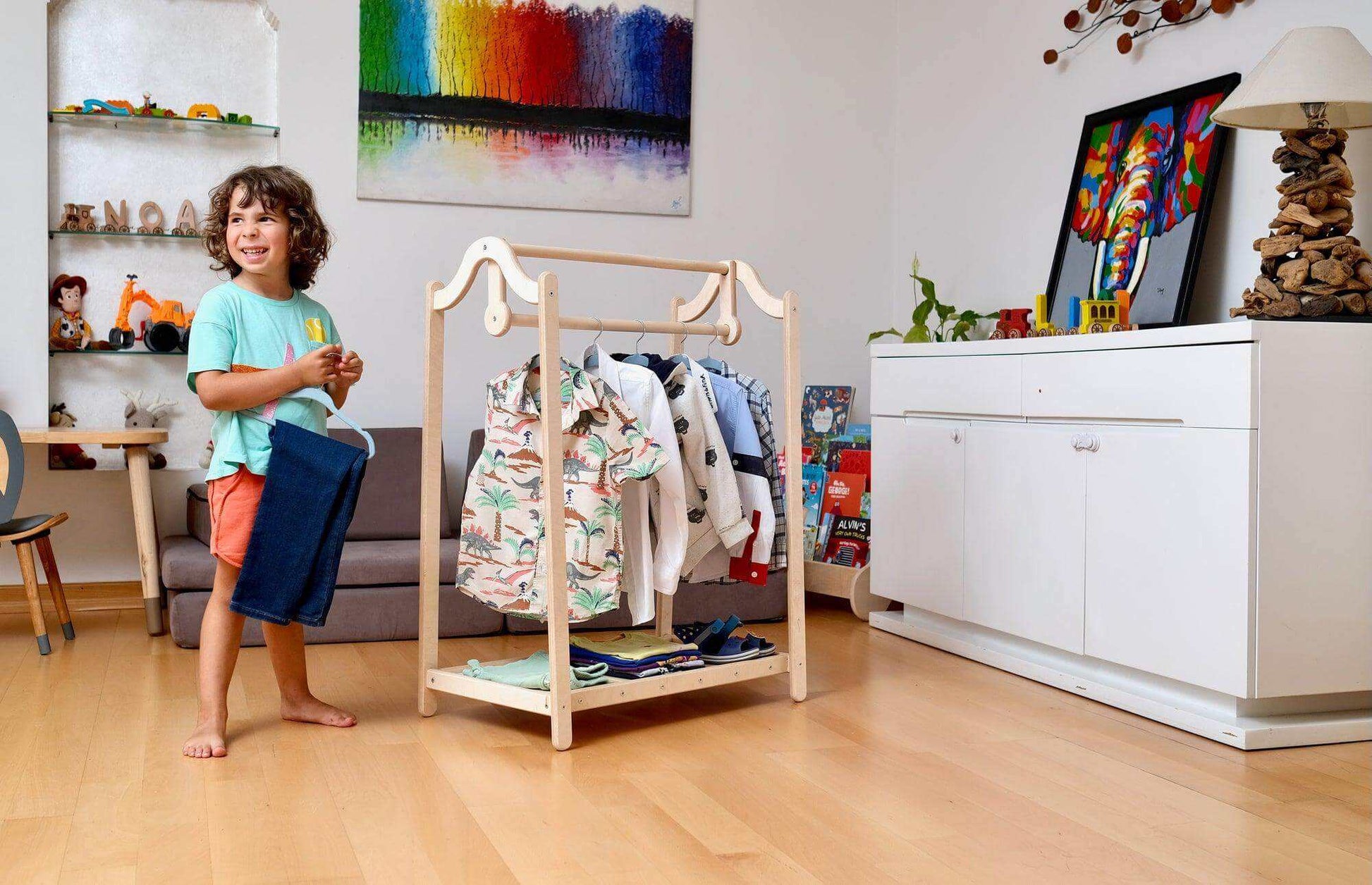 Montessori wooden kids clothing rack with adjustable hanging bar and open shelf, showing children's clothes in a modern playroom.