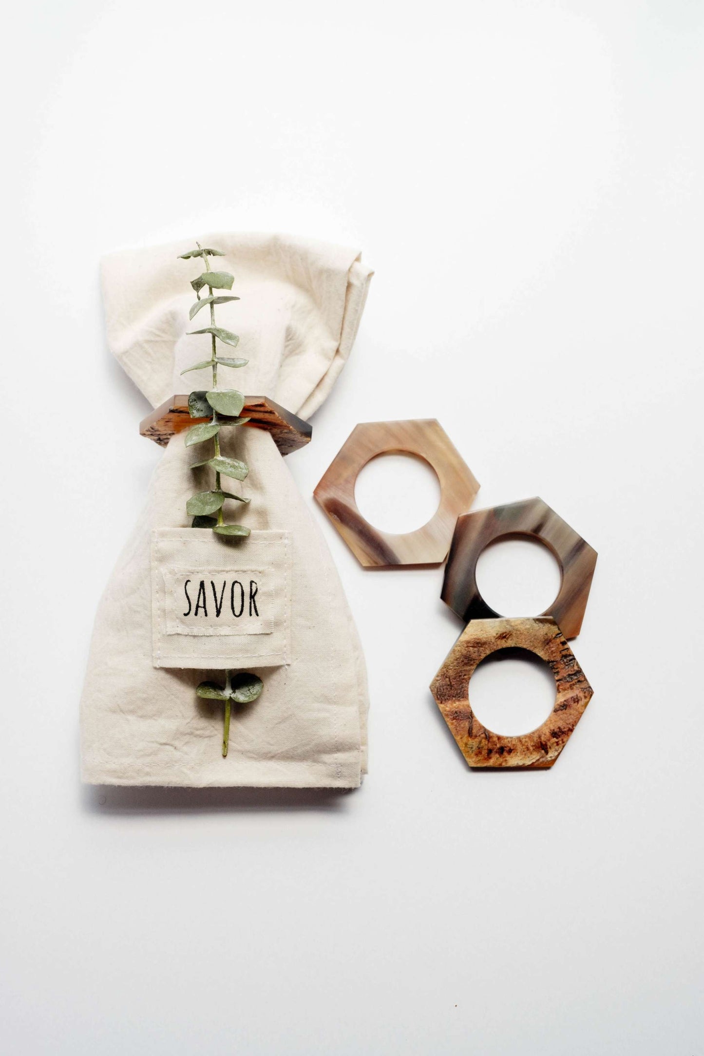 Set of 4 hexagon horn napkin rings with natural wood tones beside folded cloth napkin with greenery and savor tag