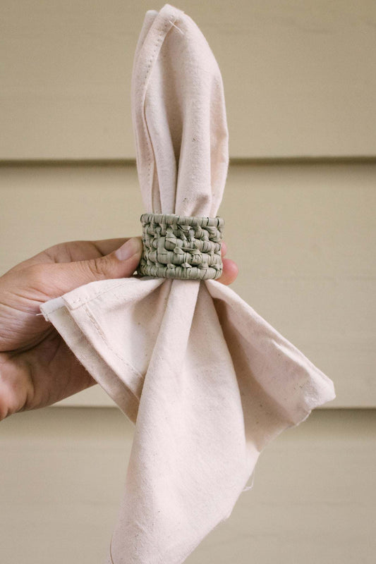 Handwoven palm leaf napkin ring holding a light beige cotton napkin against a neutral background