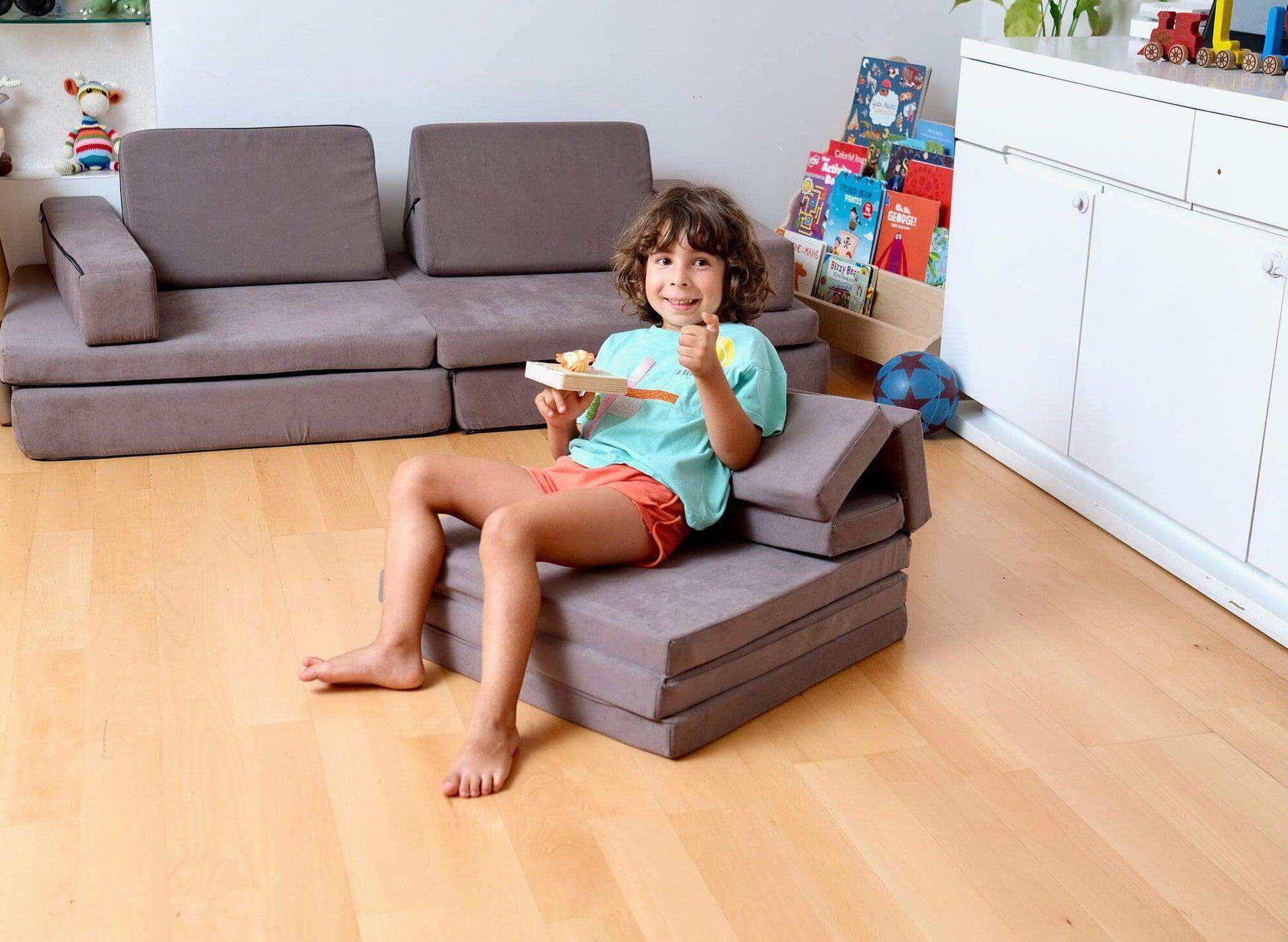 Child sitting on a gray transformable kids play mat and couch in a playroom with books and toys.