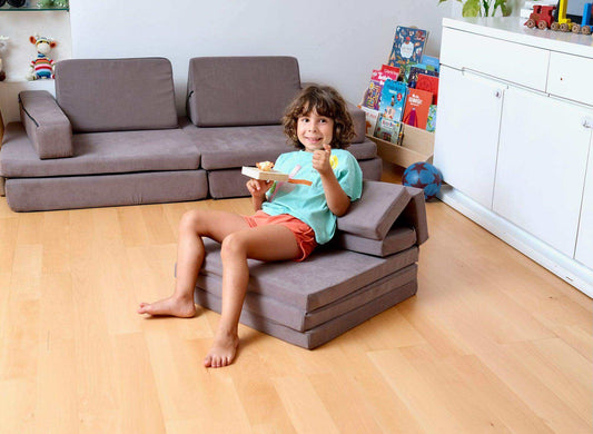Child sitting on a gray transformable kids play mat and couch in a playroom with books and toys.
