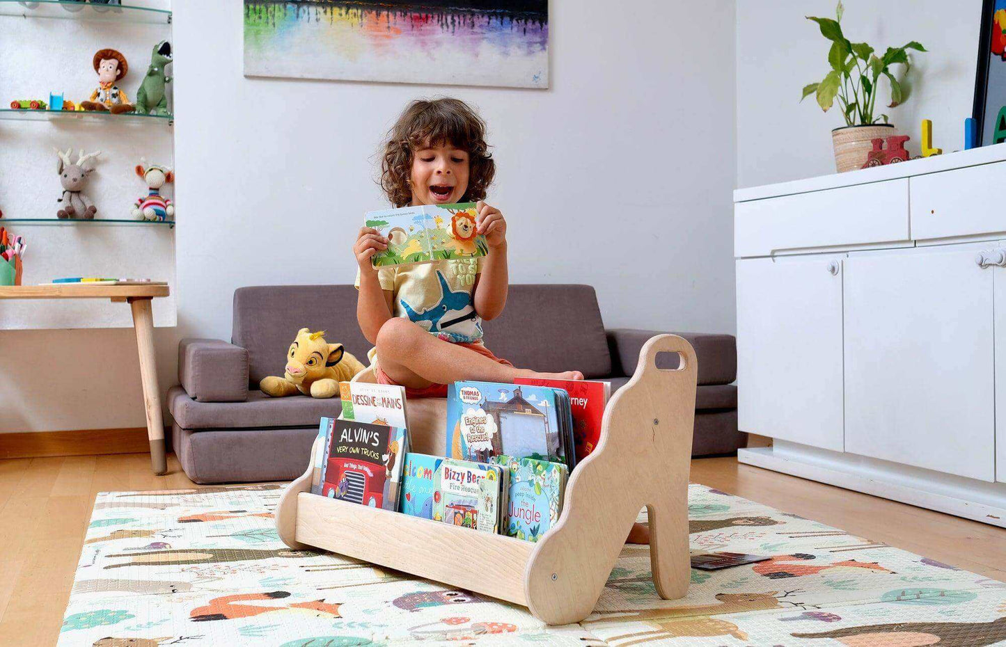 Curved wooden bookshelf with wave design holding children's books in a playroom with a child sitting behind it reading a book