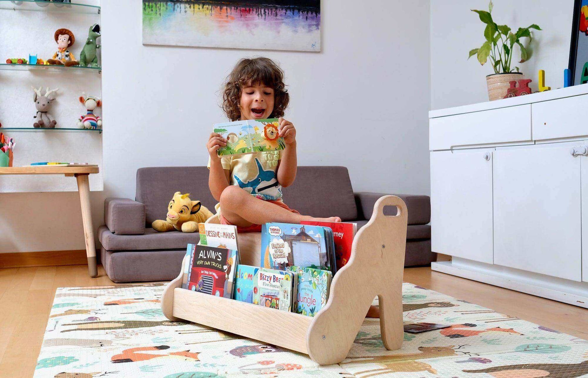 Curved wooden bookshelf with wave design holding children's books in a playroom with a child sitting behind it reading a book