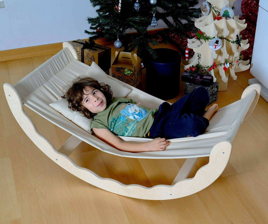 Toddler relaxing on a wooden hammock with fabric sling indoors near a decorated Christmas tree