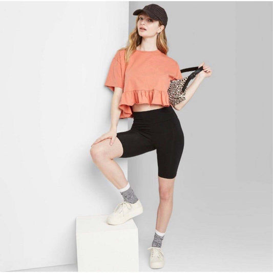 Woman wearing black high-rise bike shorts with an orange crop top and cap posing indoors